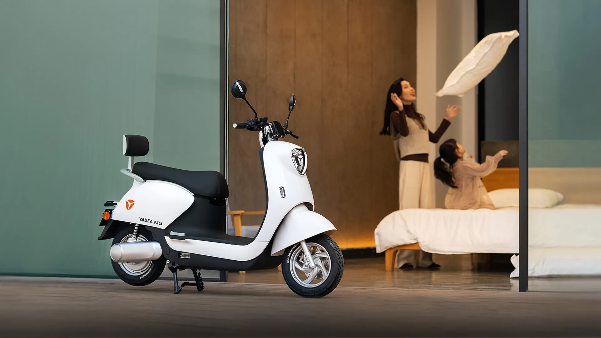 Scooter électrique rétro YADEA M6 blanc garé devant une baie vitrée, dans une chambre où une mère et sa fille jouent avec un oreiller