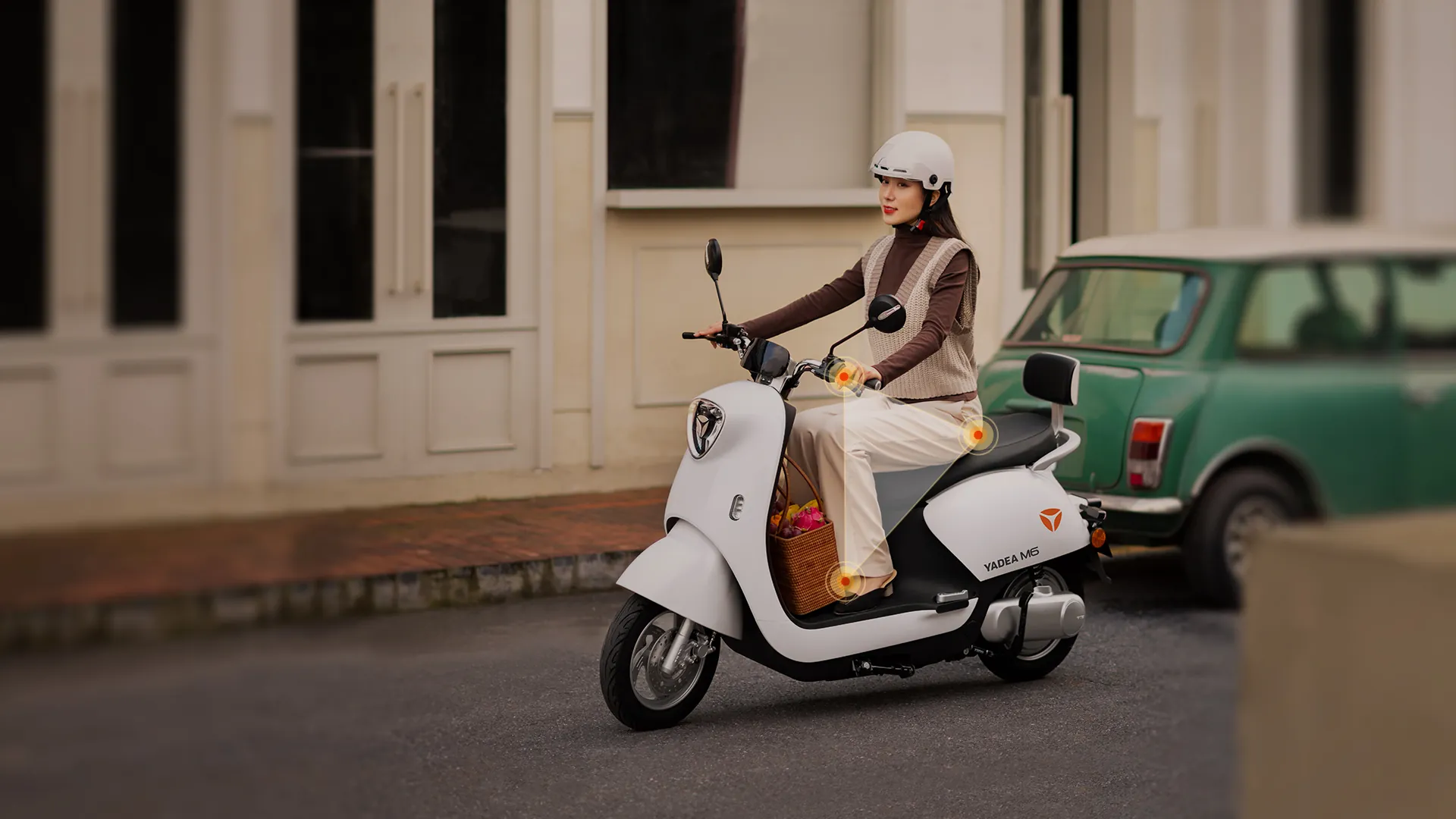 Conductrice casquée sur un YADEA M6 blanc dans une rue calme, à côté d'une voiture verte ; des points orange marquent la selle, le plancher et le guidon